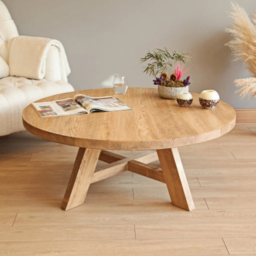 Top front view of the round coffee table showcasing its natural wood grain, sturdy legs, and a decorative arrangement, with an armchair softly blurred in the background.