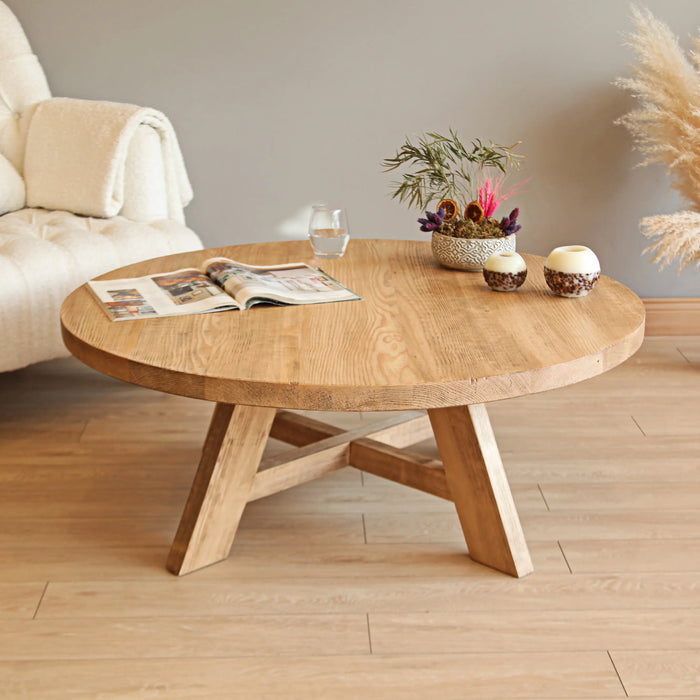 Top front view of the round coffee table showcasing its natural wood grain, sturdy legs, and a decorative arrangement, with an armchair softly blurred in the background.