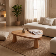A low, organic-shaped Yuma Organic Triangle Beech Wood Coffee Table with chunky legs in a warm, modern minimalist living room. The table is styled with a small vase of dried stems, a small ceramic bowl, and an open magazine, set on a large woven jute rug next to a cream boucle sofa.