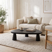 Yuma Organic Triangle Beech Wood Coffee Table with chunky legs in a bright, modern living room. The table is styled with a small stack of books and a white mug, set on a textured cream rug in front of a light beige sofa.