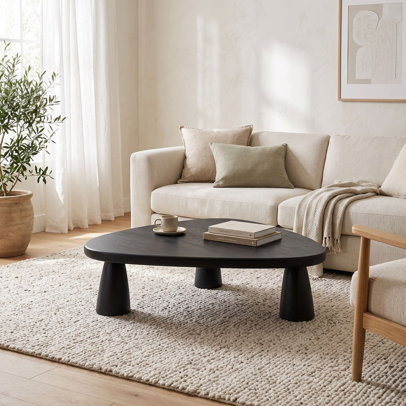 Yuma Organic Triangle Beech Wood Coffee Table with chunky legs in a bright, modern living room. The table is styled with a small stack of books and a white mug, set on a textured cream rug in front of a light beige sofa.