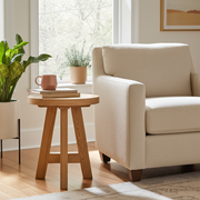 Amber Round Farmhouse Side Table
 with three legs sits next to a cream upholstered armchair in a bright living room. A pink mug and books rest on the table, with potted plants and a window in the background.