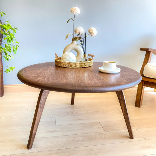 Top front view of the round coffee table showcasing its natural wood grain, sturdy legs, and a decorative arrangement, with an armchair in the background.