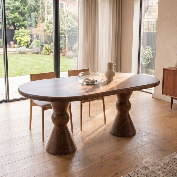 Avero Oval Solid Walnut Dining Table – Double Sculptural Pedestal Designwith chunky turned pedestal legs, shown in a bright modern room with light wood floors, dining chairs, and large windows overlooking a garden.