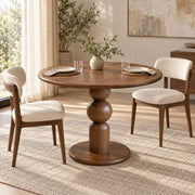 Dorel round wooden dining table with a sculptural ball-and-stick pedestal base, shown with two upholstered wooden chairs in a bright, modern dining room setting.