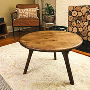 A round wooden coffee table with three tapered legs in a cozy living room setting, featuring a patterned rug, a brown leather armchair, and a wood-log wall in the background.