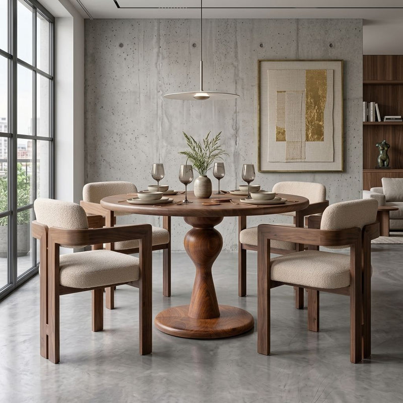 Mason Solid Walnut Round Pedestal Dining Table | Sculptural Base
 with a pedestal base surrounded by four matching wooden armchairs with light beige upholstered seats, set in a modern dining room with concrete walls and a large window.