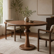 Mason Solid Walnut Round Pedestal Dining Table | Sculptural Base with a turned pedestal base and matching wooden chairs in a softly lit, neutral-toned dining room.