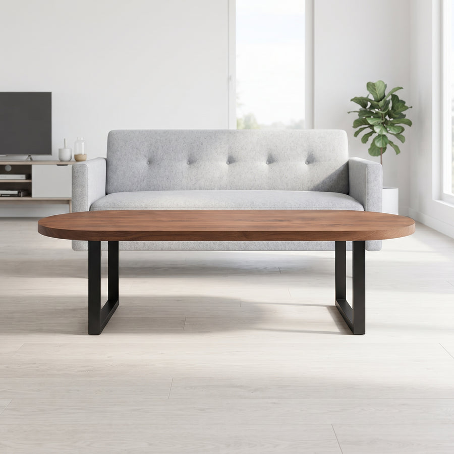 Oasis oval walnut coffee table with a medium brown wood top and black metal legs, positioned in front of a light gray tufted sofa in a bright, minimalist living room.