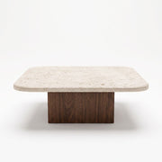 Eye-level straight-on studio shot of Pera Square Travertine Coffee Table featuring a rounded-corner beige stone top and solid walnut block base