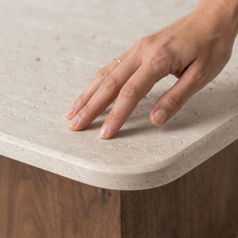 Detailed studio close-up showing human hand touching the porous texture and rounded corner of beige travertine top on Pera Square Coffee Table