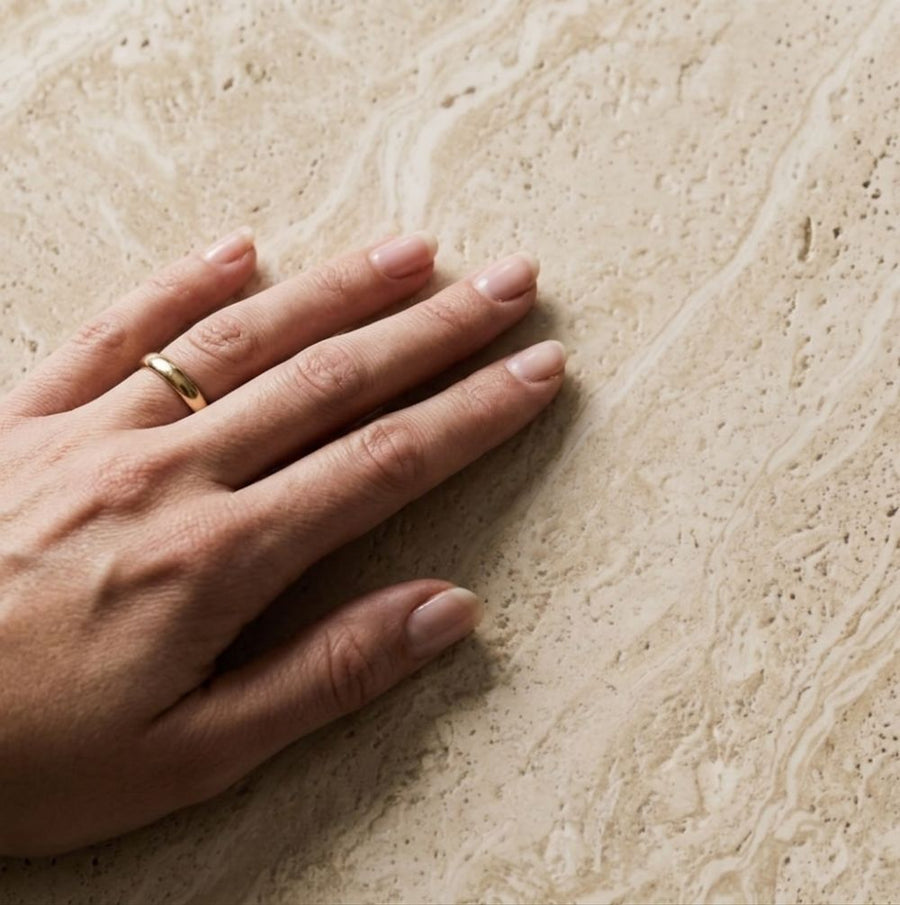 Detailed studio close-up showing human hand touching the porous texture and natural veining of beige travertine top on Pera Square Coffee Table