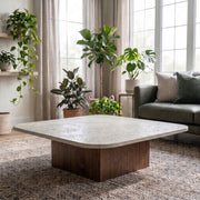 Pera Square Travertine Coffee Table with Solid Walnut Block Base Styled on Rug with Green Sofa and Indoor Houseplants in Bright Living Room