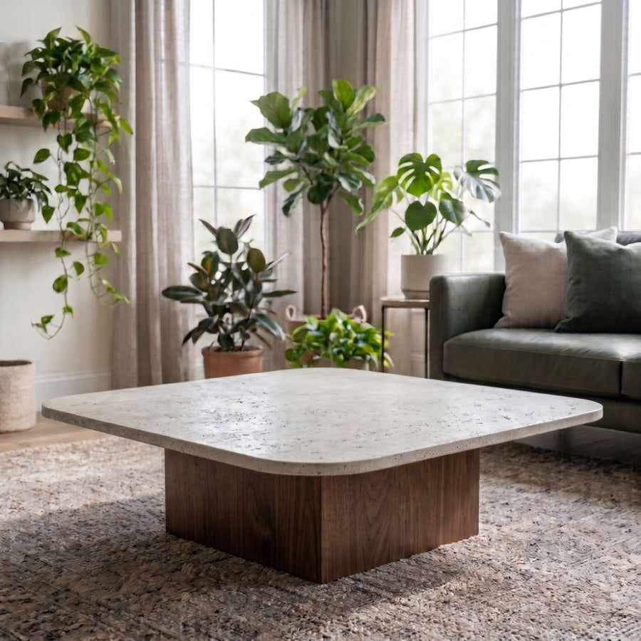 Pera Square Travertine Coffee Table with Solid Walnut Block Base Styled on Rug with Green Sofa and Indoor Houseplants in Bright Living Room