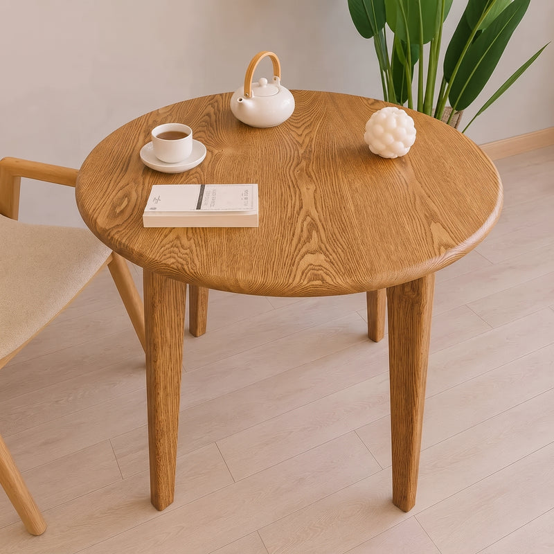 Petra Round Wood Dining Room Table Solid Oak Wood with tapered legs, styled with a white teapot, a cup of tea, a book, and a decorative object, set in a bright room with a chair and a plant.