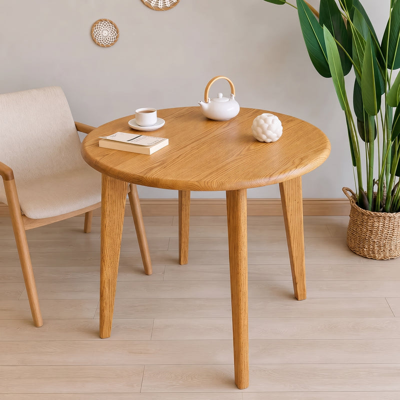 Petra Round Wood Dining Room Table Solid Oak Wood with tapered legs in a bright, modern room setting, styled with a white teapot, a book, and a cup on top.