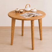 Petra Round Wood Dining Room Table Solid Oak Wood with four tapered legs, styled with a white teapot, teacup, two small ceramic vases, and an open magazine on a light wood floor against a neutral beige wall.