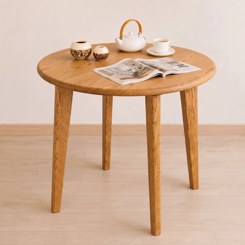 Petra Round Wood Dining Room Table Solid Oak Wood with four tapered legs, styled with a white teapot, teacup, two small ceramic vases, and an open magazine on a light wood floor against a neutral beige wall.