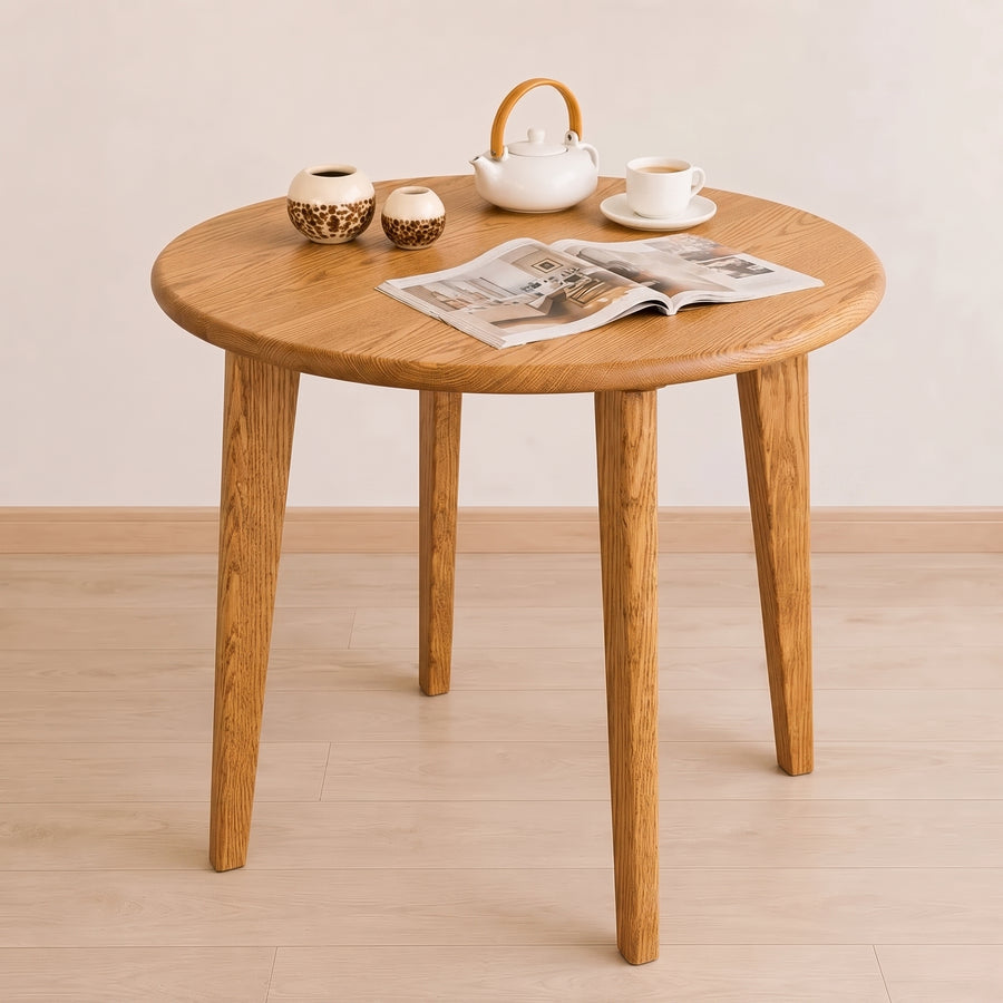 Petra Round Wood Dining Room Table Solid Oak Wood with four tapered legs, styled with a white teapot, teacup, two small ceramic vases, and an open magazine on a light wood floor against a neutral beige wall.
