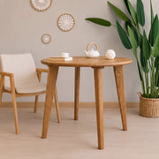 Petra Round Wood Dining Room Table Solid Oak Wood with tapered legs, styled in a minimalist room with a cream upholstered wooden chair, a white teapot, and a potted green plant.