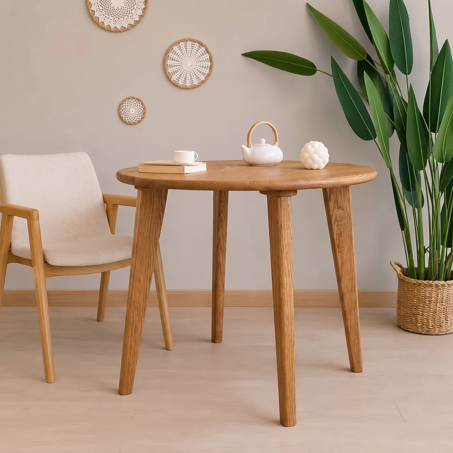 Petra Round Wood Dining Room Table Solid Oak Wood with tapered legs, styled in a minimalist room with a cream upholstered wooden chair, a white teapot, and a potted green plant.
