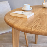 A close-up of Petra Round Wood Dining Room Table Solid Oak Wood with a rounded edge and tapered legs, styled with a book, a cup of coffee, and a teapot in a bright, modern room.
