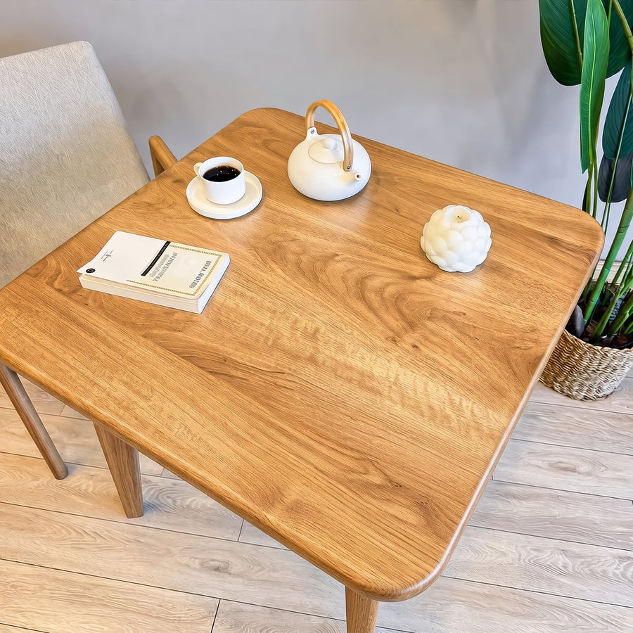 Petra Square Wood Dining Table Solid Oak Wood with rounded corners and a warm oak finish, styled in a bright room with a white teapot, a cup of coffee, a book, and a potted plant.