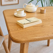 Petra Square Wood Dining Table Solid Oak Wood in a bright room, styled with a white teapot, a cup and saucer, a candle, and a book in the foreground.