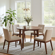 Petra Round Wood Dining Room Table | Walnut Wood Mid Century Design  featuring a round wooden table and four upholstered chairs with beige fabric and dark wood legs, arranged on a patterned rug in a bright dining room.