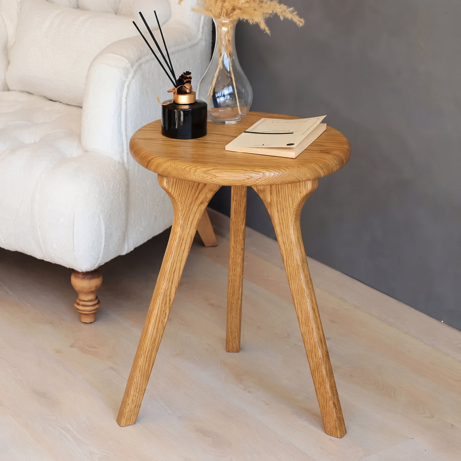 Sidney Round Side Table Solid Oak Woodwith three angled legs, styled in a living room next to a white tufted armchair. 
