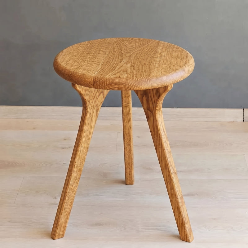Sidney Round Side Table Solid Oak Wood with three splayed legs, made from light oak wood with a visible grain pattern, set against a gray wall and light wood floor.