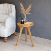 Sidney Round Side Table Solid Oak Wood with three angled legs, styled with a glass vase of pampas grass, a black reed diffuser, and a notebook, next to a white tufted armchair against a dark gray wall.