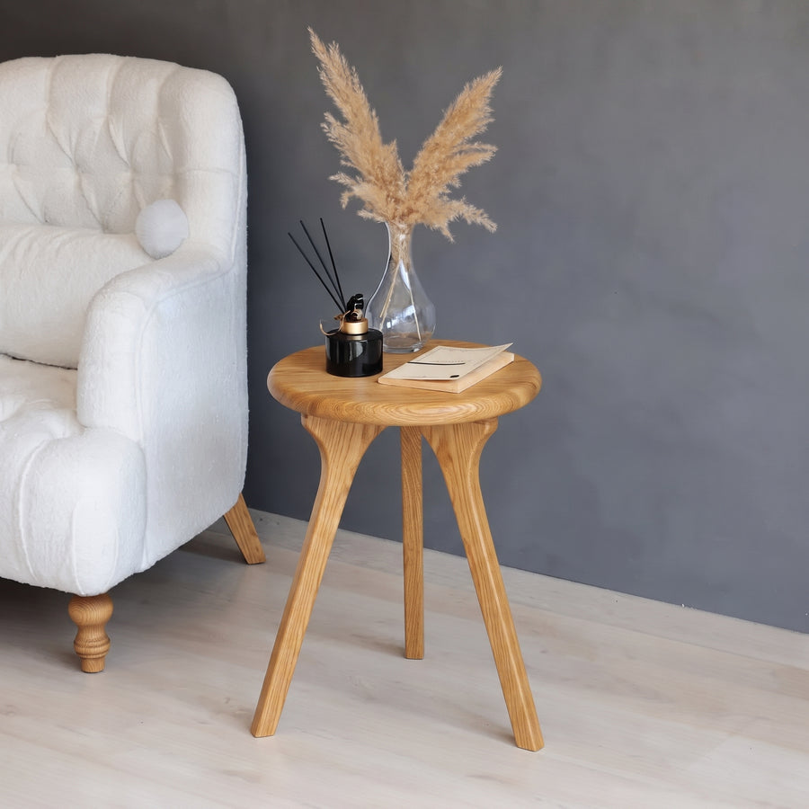 Sidney Round Side Table Solid Oak Wood with three angled legs, styled with a glass vase of pampas grass, a black reed diffuser, and a notebook, next to a white tufted armchair against a dark gray wall.