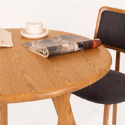 Sidney Circular Dining Table Solid Oak Wood – Mid-Century Modern Design with a warm oak finish, styled with a folded magazine and a white cup and saucer, next to a dark gray upholstered wooden chair on a white background.