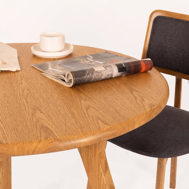Sidney Circular Dining Table Solid Oak Wood – Mid-Century Modern Design with a warm oak finish, styled with a folded magazine and a white cup and saucer, next to a dark gray upholstered wooden chair on a white background.