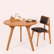 Sidney Circular Dining Table Solid Oak Wood – Mid-Century Modern Design with three legs and a matching wooden chair with dark upholstered seat and backrest, styled on a clean off-white background with small tabletop props.