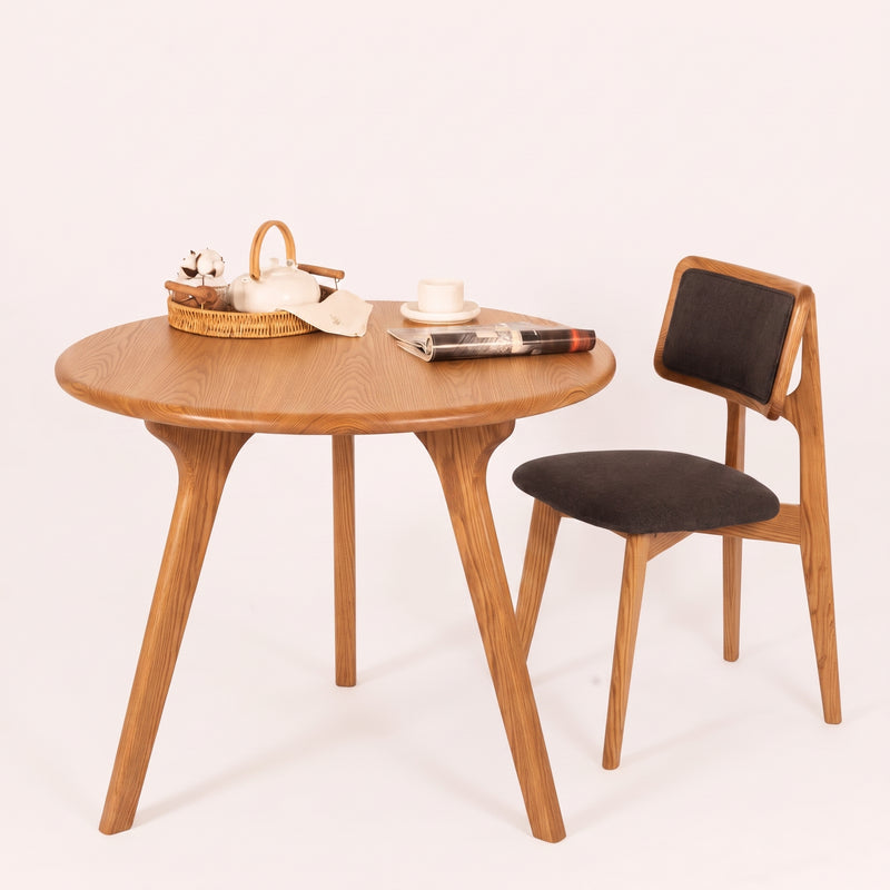 Sidney Circular Dining Table Solid Oak Wood – Mid-Century Modern Design with three legs and a matching wooden chair with dark upholstered seat and backrest, styled on a clean off-white background with small tabletop props.