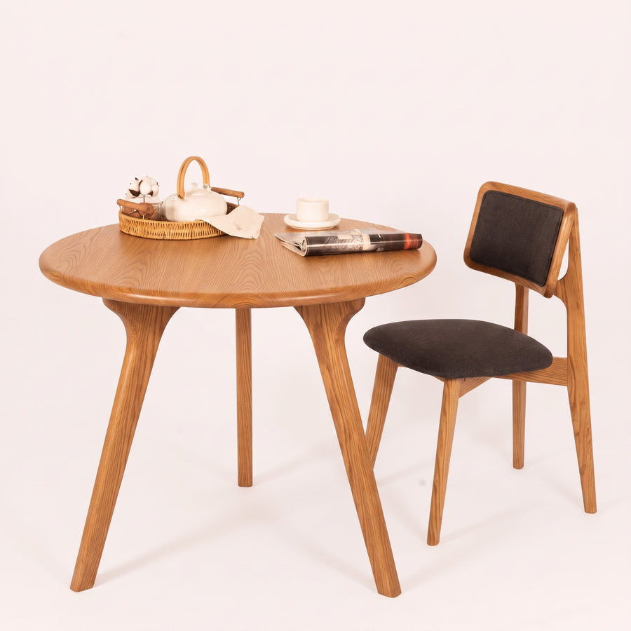 Sidney Circular Dining Table Solid Oak Wood – Mid-Century Modern Design with three legs and a matching wooden chair with dark upholstered seat and backrest, styled on a clean off-white background with small tabletop props.