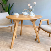 Sidney Circular Dining Table Solid Oak Wood – Mid-Century Modern Design with three curved legs, styled with a white teapot, two white teacups, and a vase of flowers, set in a room with light wood floors and neutral walls.