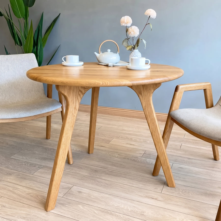 Sidney Circular Dining Table Solid Oak Wood – Mid-Century Modern Design with three curved legs, styled with a white teapot, two white teacups, and a vase of flowers, set in a room with light wood floors and neutral walls.