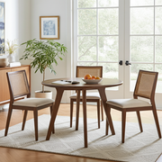 Sidney Circular Walnut Dining Table | Mid Century Modern  with three matching chairs featuring woven cane backs and cream upholstered seats, styled in a bright, modern dining room with large windows and a potted plant.