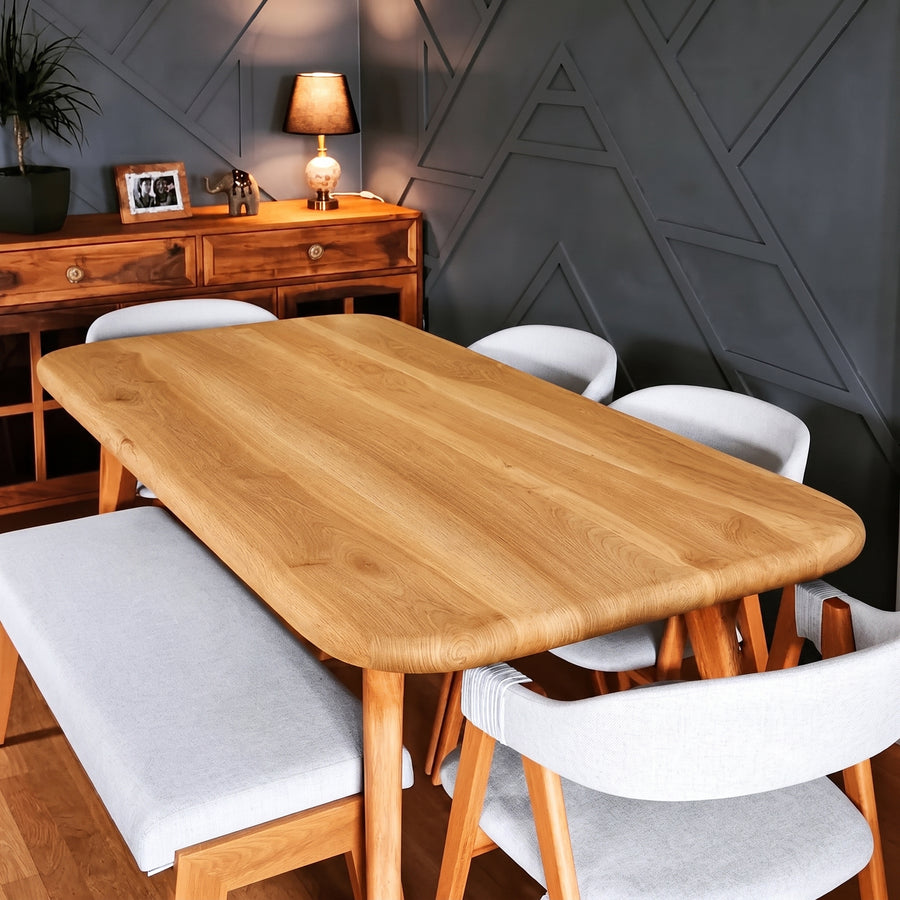 Sidney Rectangle Dining Table Solid Oak Wood – Mid-Century Modern Design with rounded corners and light wood legs, surrounded by white upholstered chairs in a styled dining room with a dark geometric wall and wooden sideboard.