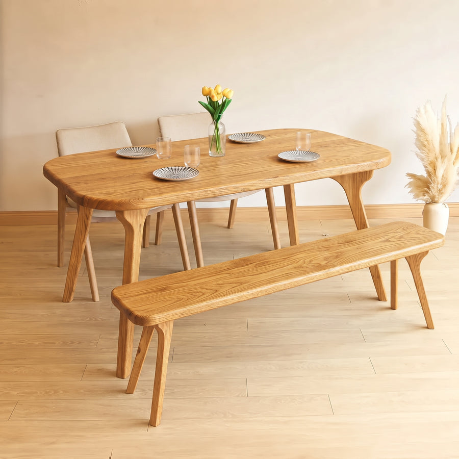 A light oak dining set featuring Sidney Rectangle Dining Table Solid Oak Wood – Mid-Century Modern Design, a matching bench, and three chairs, styled with plates, a vase of yellow tulips, and pampas grass in a minimalist room.
