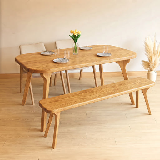 A light oak dining set featuring Sidney Rectangle Dining Table Solid Oak Wood – Mid-Century Modern Design, a matching bench, and three chairs, styled with plates, a vase of yellow tulips, and pampas grass in a minimalist room.