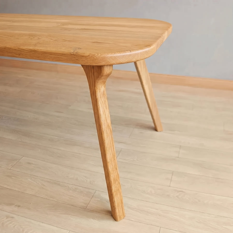 A close-up of Sidney Rectangle Dining Table Solid Oak Wood – Mid-Century Modern Design showing the rounded top edge and one of its tapered legs, set on a light wood floor against a neutral gray wall.