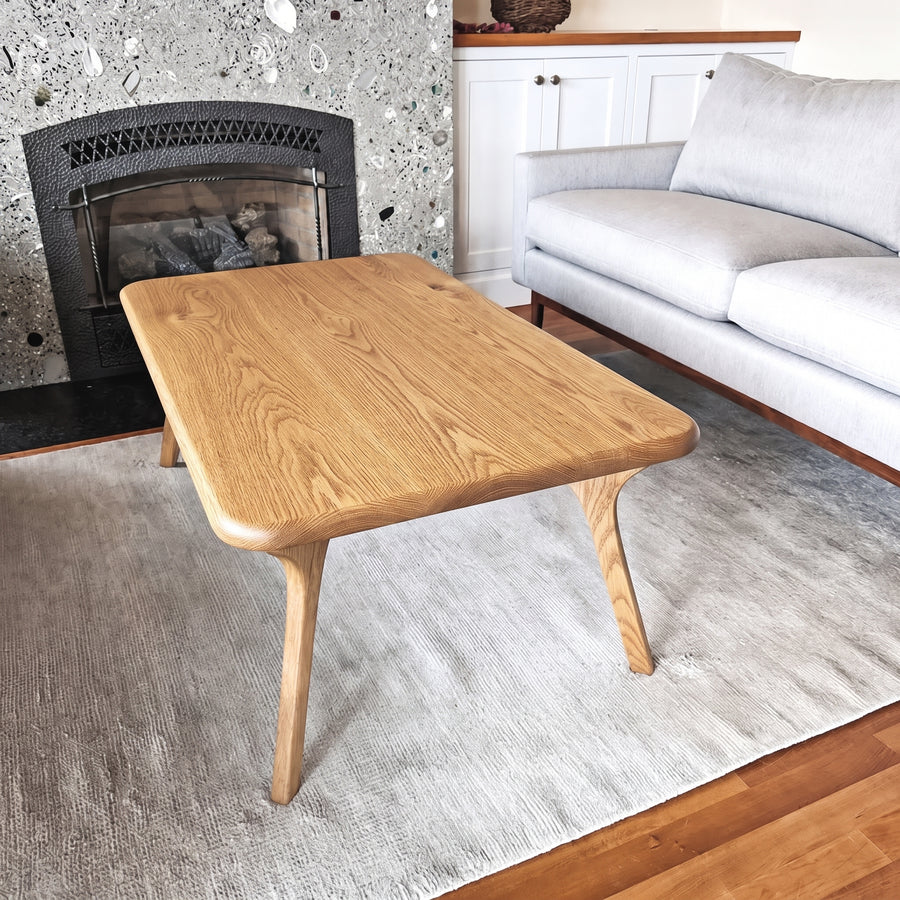 Sidney Rectangular Coffee Table Solid Oak Wood – Mid-Century Modern Design with rounded corners and tapered legs, styled in a living room setting on a gray rug next to a white sofa and a stone fireplace.