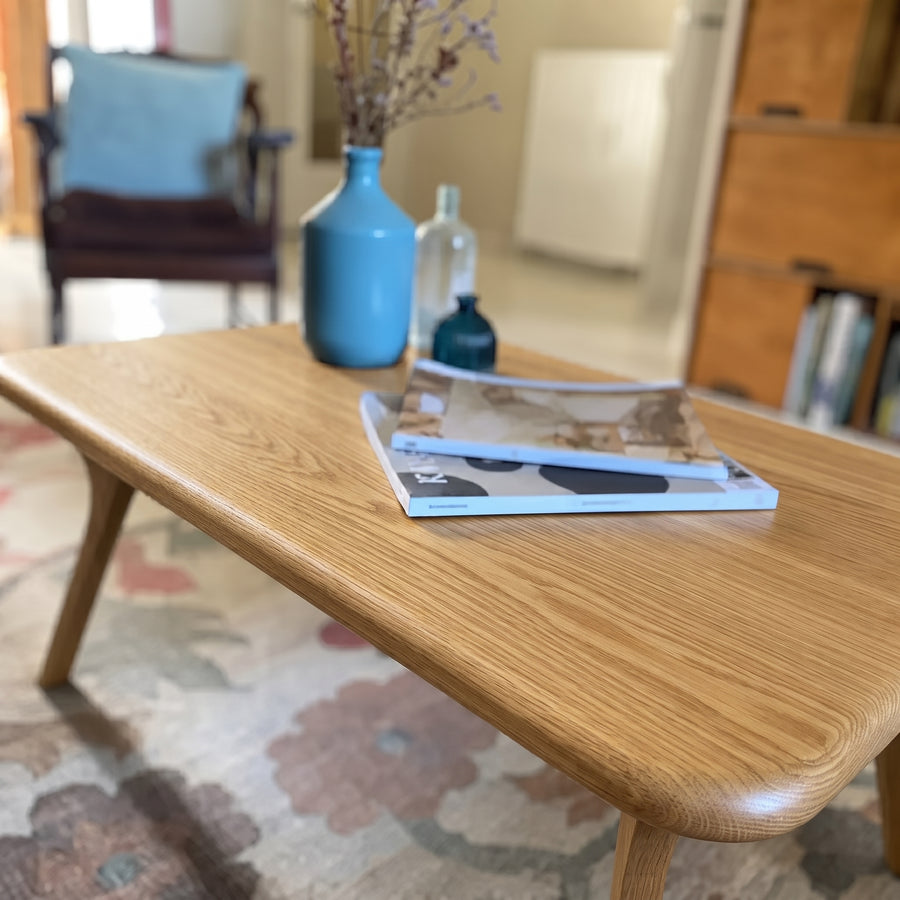 Sidney Rectangular Coffee Table Solid Oak Wood – Mid-Century Modern Design with rounded corners and tapered legs, styled with a blue ceramic vase, a clear glass bottle, and magazines in a cozy living room setting.