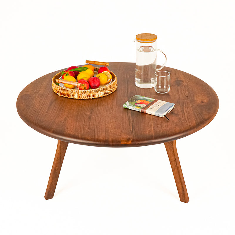 Sidney Round Coffee Table - Solid Walnut Wood - Mid Century Modern Design with tapered legs, styled with a wicker basket of fruit, a glass carafe with a wooden lid, a small glass, and a magazine on a white background.