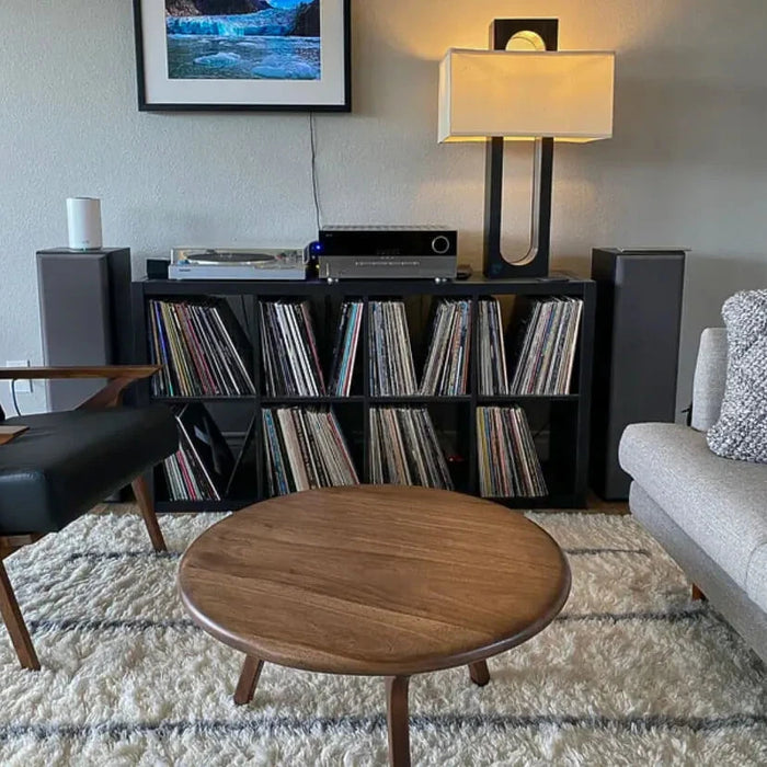 Customer photo featuring the Sidney Solid Wood Mid-Century Round Coffee Table positioned in the center of a stylish living room, flanked by a grey couch on the right and a black armchair on the left, with a black low bookcase in the background, showcasing its versatility with cool color palettes.