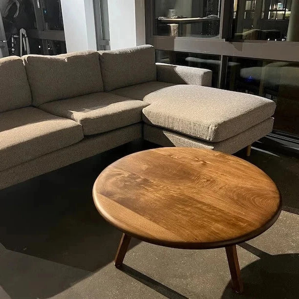 Customer photo of the Sidney Solid Wood Mid-Century Round Coffee Table placed in front of a grey couch, illuminated by direct sunlight that enhances the table's rich wood grain and creates a warm, inviting atmosphere.